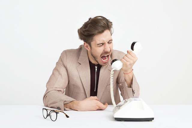 A man in suit yelling at the telephone receiver in his left hand, while his spectacles lay on the table